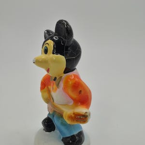 May include: A vintage figurine of a cartoon mouse character. The mouse has a black head with cracked paint, a yellow face, and is wearing an orange and pink shirt, blue pants, and black shoes. The figurine is standing on a white base.
