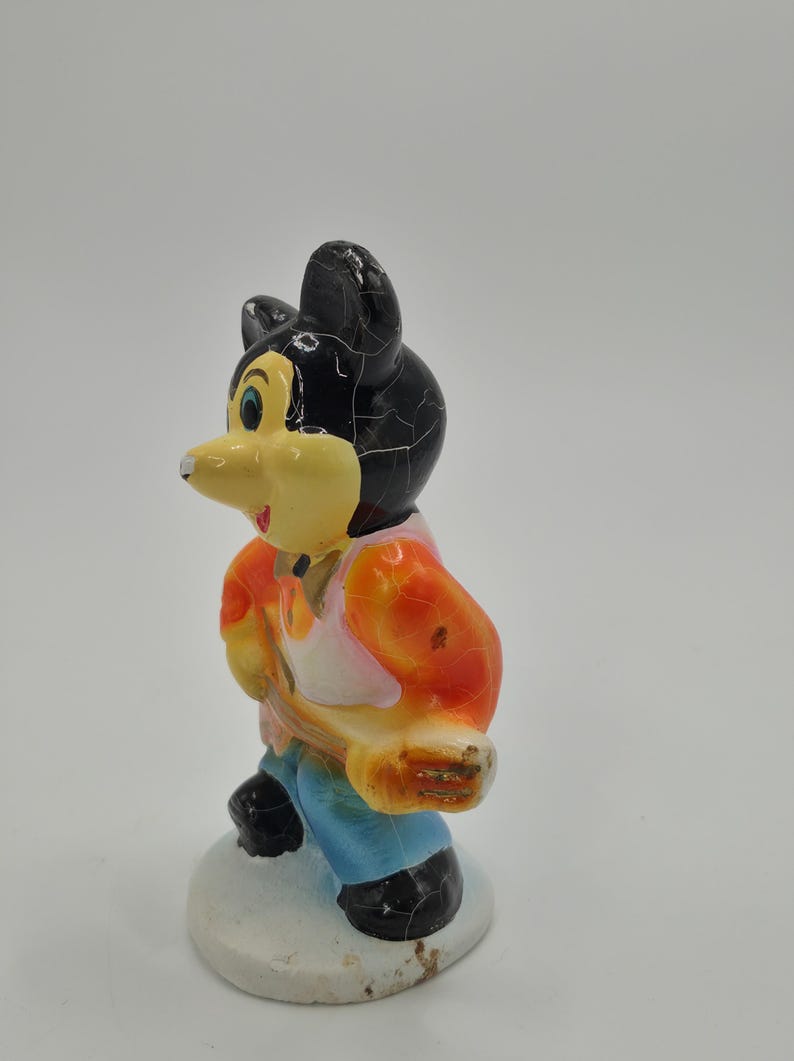 May include: A vintage figurine of a cartoon mouse character. The mouse has a black head with cracked paint, a yellow face, and is wearing an orange and pink shirt, blue pants, and black shoes. The figurine is standing on a white base.