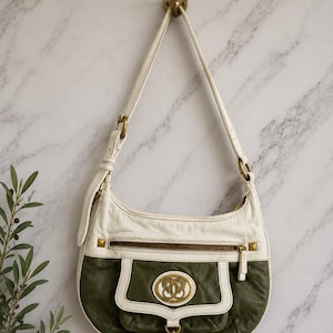 May include: A hobo-style shoulder bag featuring a two-tone design with white and olive green leather. The bag includes a zippered top pocket, a front flap pocket with a central gold-toned circular emblem, and antique brass hardware accents.
