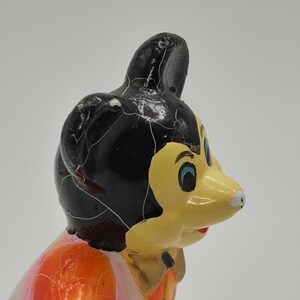 May include: A vintage figurine of a cartoon character with a black head, yellow face, and orange and pink clothing. The figurine has a cracked, aged appearance, suggesting it is an antique or collectible item.
