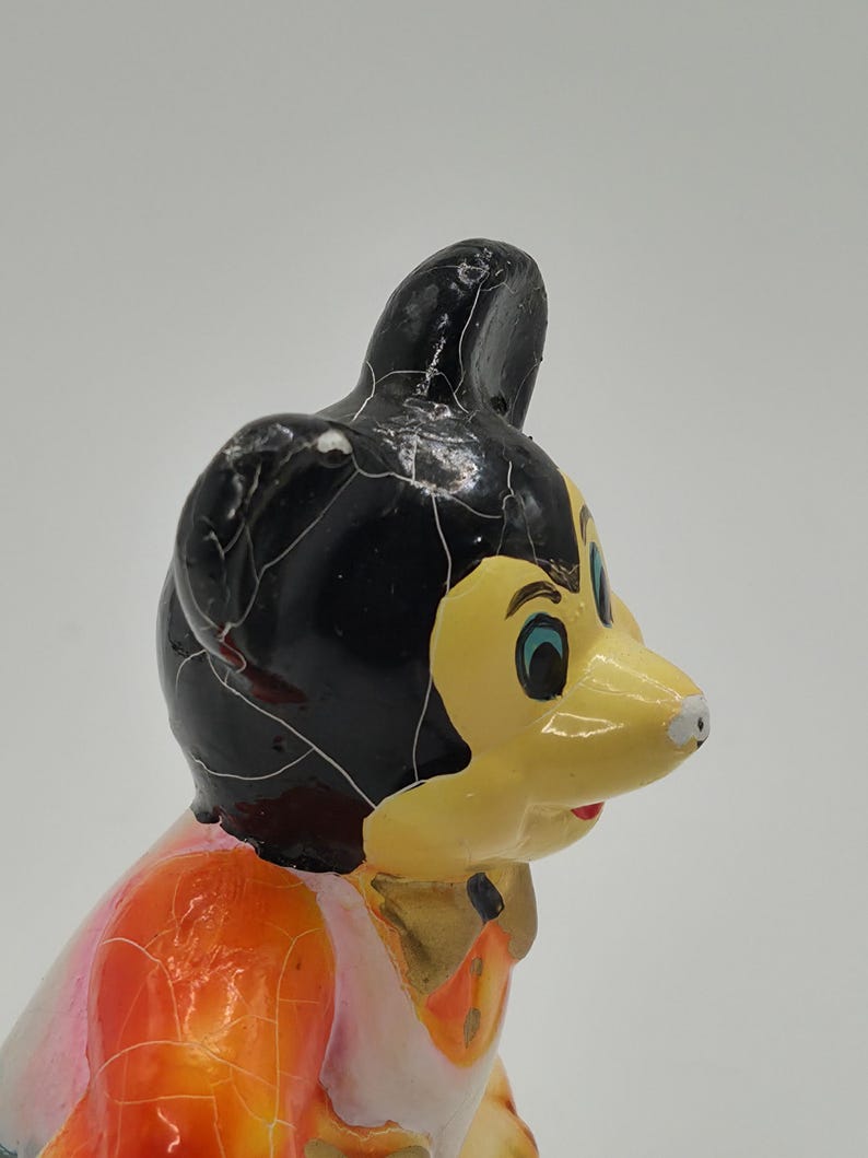 May include: A vintage figurine of a cartoon character with a black head, yellow face, and orange and pink clothing. The figurine has a cracked, aged appearance, suggesting it is an antique or collectible item.