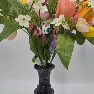 May include: A floral arrangement featuring colorful tulips, white flowers, and green leaves in a dark gray marble-look vase. The tulips are in shades of pink, orange, and yellow. The vase has a classic, elegant shape.