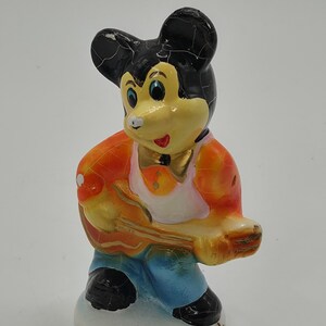 May include: A vintage figurine of a cartoon mouse playing a guitar. The mouse has a black head, yellow face, and is wearing an orange jacket, blue pants, and black shoes. The figurine is on a white base.
