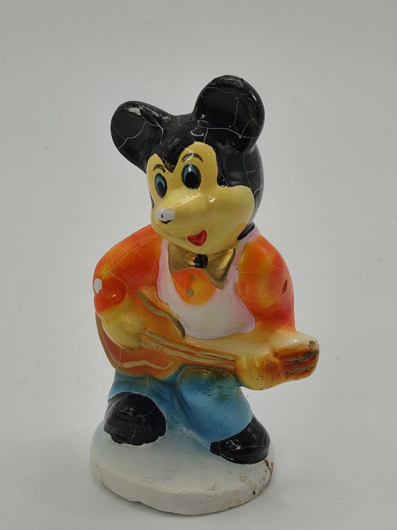 May include: A vintage figurine of a cartoon mouse playing a guitar. The mouse has a black head, yellow face, and is wearing an orange jacket, blue pants, and black shoes. The figurine is on a white base.