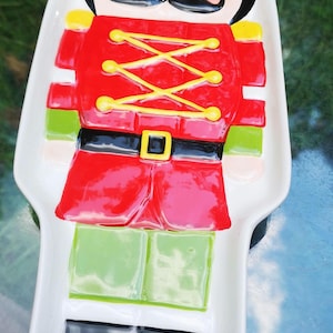 May include: A white ceramic serving platter in the shape of a toy soldier. The soldier is depicted in red, green, black, and yellow, with a black mustache and a yellow crown. The platter is approximately 12 inches long.