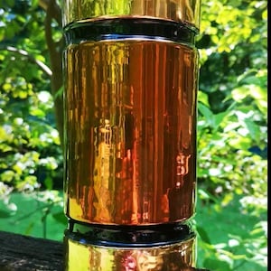 May include: A cylindrical jar with a shiny, metallic gold finish. The jar has a matching gold lid and base, with a black band separating the gold sections. The jar is set against a backdrop of green foliage.