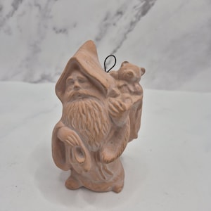 May include: A terracotta-colored clay ornament featuring a bearded wizard wearing a pointed hat and robes, carrying a small bear on their back. The figure has a metal wire loop at the top for hanging and displays detailed sculpted facial features.