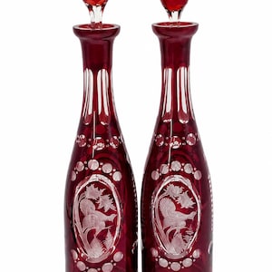 May include: A pair of tall, ruby red glass decanters with matching stoppers. Each decanter features clear cut-glass details, including vertical fluting at the neck and an oval medallion on the body depicting a bird perched among foliage.