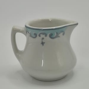 May include: A small, white ceramic creamer with a curved handle and spout. The rim is accented with a light blue and gray decorative pattern. The creamer has a rounded body and is suitable for serving cream or milk.