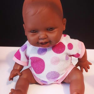 May include: A brown baby doll with brown eyes and a pink and white polka dot onesie.
