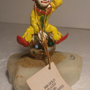 May include: A hand-painted, 24K gold-plated clown figurine balancing on a polka-dotted ball. The clown wears a yellow suit, red shoes, and a gold hat. The figurine sits on a star-shaped base with gold beads. A tag reads "24K GOLD PLATED AND HAND PAINTED."