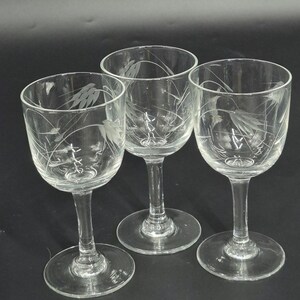 May include: Three clear, stemmed wine glasses with etched floral designs. The glasses have a rounded bowl, a slender stem, and a circular base. The etched design features delicate leaves and stems. The glasses are arranged on a dark surface.