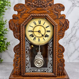 May include: A wooden mantel clock with a dark brown finish and intricate carved scrollwork. The clock face features a cream-colored dial with black Roman numerals and hands. A silver-toned pendulum hangs behind a glass panel with white floral details.
