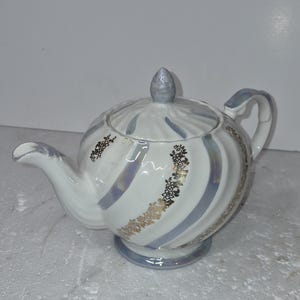 May include: White ceramic teapot with a rounded body, adorned with blue and gold floral accents. The teapot features a curved spout, a handle, and a lid with a small knob. The design includes blue stripes and gold floral patterns.
