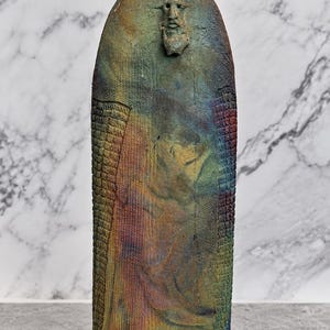 May include: A tall, cylindrical ceramic vase with a flared, wide rim. The surface features a textured, multi-colored finish in shades of blue, green, and bronze. A small, sculpted human face is centered near the top of the vessel body.