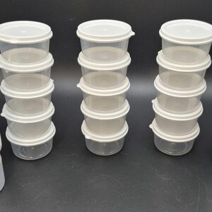 May include: A collection of small, clear plastic containers with white lids. The containers are stacked in multiple columns, with a few individual containers visible. The items are likely for storage or organization.