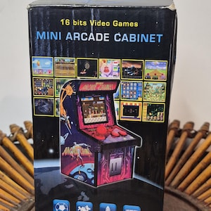 May include: A black box featuring a mini arcade cabinet with the text "16 bits Video Games MINI ARCADE CABINET." The box has game graphics and icons for sport, puzzle, arcade, and shooting games. The arcade cabinet is red and purple.