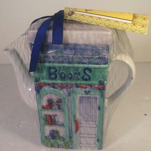 May include: A ceramic teapot with a shop-front design, wrapped in clear plastic. The shop front is green and blue with the word "Books" above the door. A blue ribbon and a yellow tag are attached.