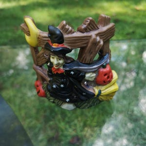 May include: A Halloween-themed ceramic figurine featuring a witch riding a broom. The witch is dressed in a black outfit with a pointed hat and holds a carved pumpkin. The figurine is set against a brown fence and a yellow crescent moon.