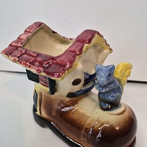 May include: A ceramic boot-shaped planter with a house-like roof and a blue squirrel figurine. The boot is brown, tan, and black, with a red roof and yellow accents. The squirrel has a blue body and yellow tail.