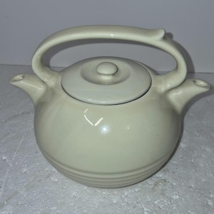 May include: Cream-colored ceramic teapot with a rounded body and a curved handle. The teapot has a lid with a small knob and two spouts. The body features horizontal ridges near the base.