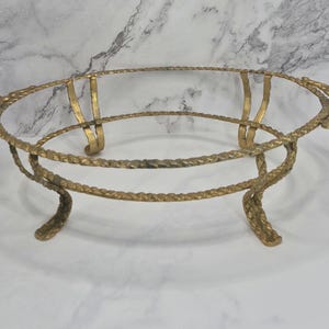 May include: An oval-shaped gold-toned metal stand featuring a twisted rope-like frame design. The stand is supported by four curved legs and includes two decorative shell-shaped handles on opposite ends, providing a stable base for a dish.