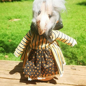 May include: A handmade Halloween doll with a witch-like appearance. The doll has a fabric face with a long nose and gray hair. It wears a striped orange and black dress with a black skirt featuring orange stars. The doll is standing on a wooden surface.
