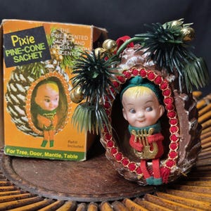May include: A vintage Christmas ornament shaped like a pine cone, featuring a pixie doll holding a harp. The ornament is decorated with red and green accents, and the original box is visible, with the text "Pixie Pine-Cone Sachet".