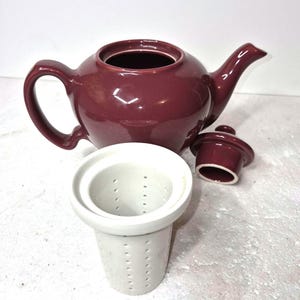 May include: A burgundy ceramic teapot with a matching lid and a removable white tea infuser. The teapot has a rounded body, a curved spout, and a large handle. The infuser is cylindrical with small holes.