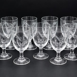 May include: A set of nine clear wine glasses. Each glass has a stem and a bowl with an etched wheat design. The glasses are arranged on a black surface.