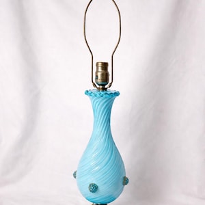 May include: A vintage blue glass table lamp with a swirled design. The lamp has a round base, a bulbous body, and a brass neck and lamp holder. Decorative blue accents are on the body. The lamp is approximately 18 inches tall.