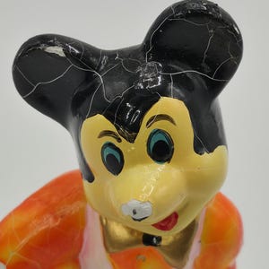May include: Vintage Mickey Mouse figurine with black ears, a yellow face, and a red mouth. The figurine has a gold bow tie and an orange and white outfit. The paint shows signs of age and wear, with visible cracks.