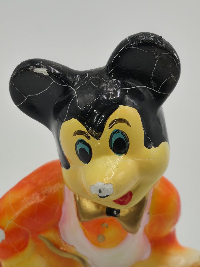 May include: Vintage Mickey Mouse figurine with black ears, a yellow face, and a red mouth. The figurine has a gold bow tie and an orange and white outfit. The paint shows signs of age and wear, with visible cracks.
