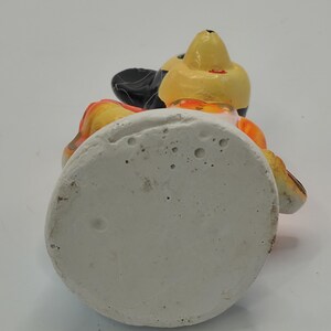 May include: A ceramic figurine of a cartoon character, featuring a yellow head, black ears, and orange and black accents. The figurine is set on a white, circular base. The character is wearing a black hat.