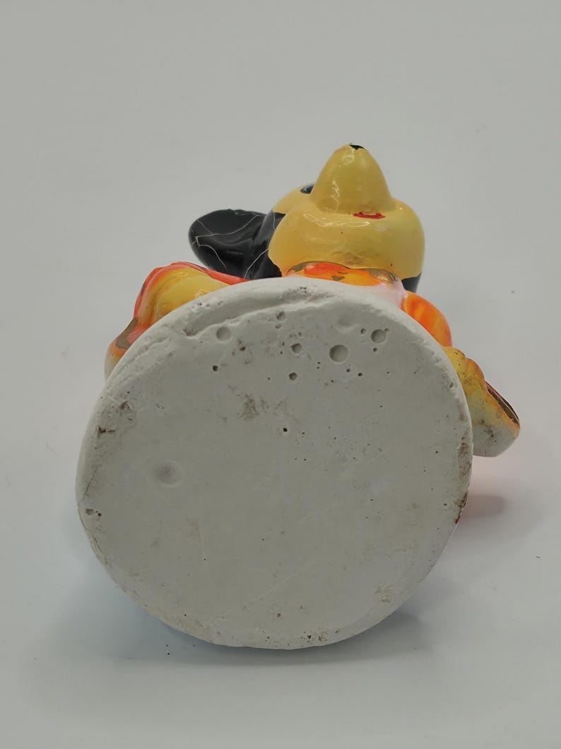 May include: A ceramic figurine of a cartoon character, featuring a yellow head, black ears, and orange and black accents. The figurine is set on a white, circular base. The character is wearing a black hat.