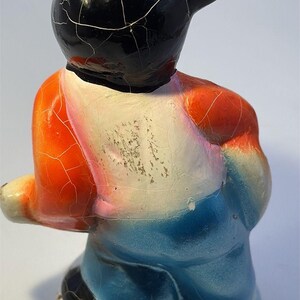 May include: A vintage ceramic figurine of a bear-like character, featuring a black head with a cracked glaze, orange and blue clothing, and a white chest. The figurine has a seated pose and a glossy finish.