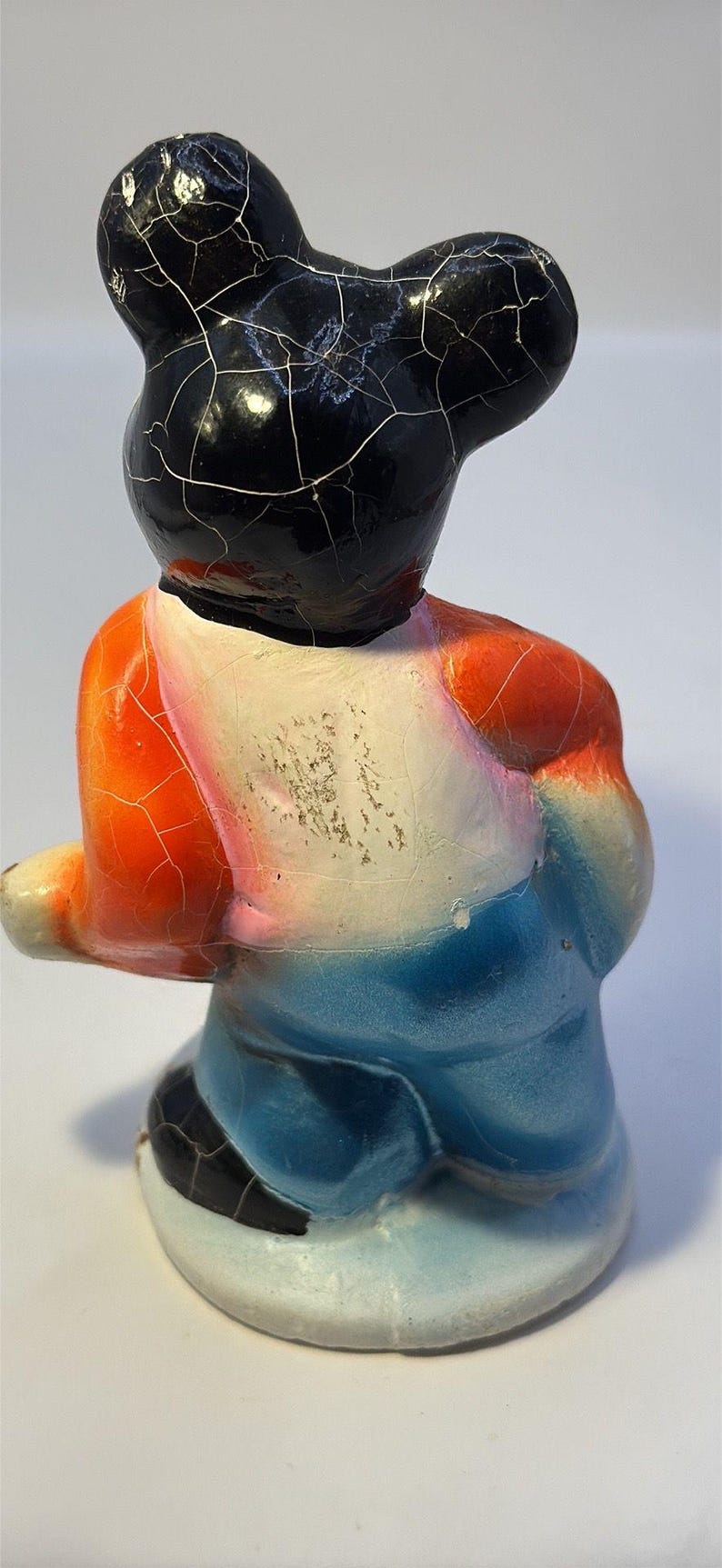 May include: A vintage ceramic figurine of a bear-like character, featuring a black head with a cracked glaze, orange and blue clothing, and a white chest. The figurine has a seated pose and a glossy finish.