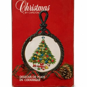 May include: A rectangular red box labeled Christmas by Carlton and Dessous de Plats en Ceramique. The box features a round ceramic trivet with a black metal frame and a decorative handle, displaying a Christmas tree with ornaments and gifts.