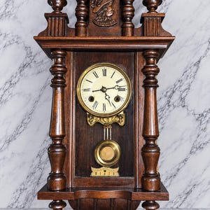May include: A dark brown wooden wall clock featuring a round white face with black Roman numerals, ornate hands, and a brass pendulum. The clock is decorated with carved pillars and a relief carving of a profile portrait at the top center.