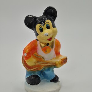May include: A vintage figurine of a cartoon mouse playing a guitar. The mouse has black ears, a yellow face, and is wearing an orange jacket, blue pants, and a gold bow tie. The figurine is standing on a white base.