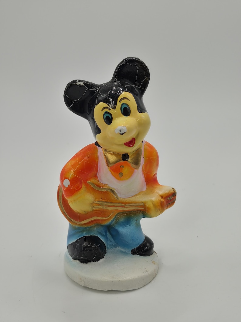 May include: A vintage figurine of a cartoon mouse playing a guitar. The mouse has black ears, a yellow face, and is wearing an orange jacket, blue pants, and a gold bow tie. The figurine is standing on a white base.