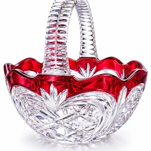 May include: A clear crystal basket with a red rim and handle. The basket features intricate cut glass patterns and a scalloped edge. The handle is arched and also cut with a geometric design. This decorative basket is ideal for display or as a gift.