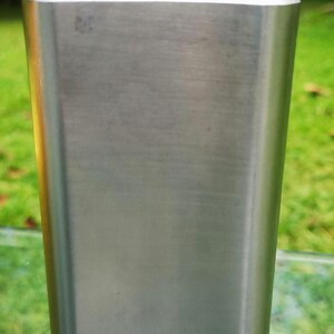 May include: A stainless steel flask with a brushed metal finish. The flask has a rectangular body with rounded edges and a hinged, screw-on cap. The cap and hinge are a shiny, gold-colored metal. The flask is designed to hold liquids.