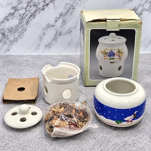 May include: A ceramic potpourri burner set including a white base with circular cutouts, a matching lid with four holes, a blue bowl with a white goose and snowflake pattern, a cardboard insert, and a plastic bag filled with dried potpourri.