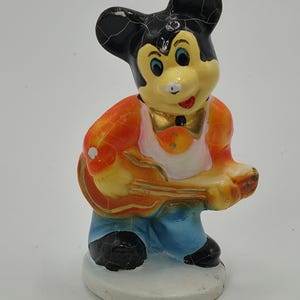 May include: A vintage figurine of a cartoon mouse playing a guitar. The mouse has a yellow face, black ears, and is wearing an orange vest, blue pants, and black shoes. The guitar is yellow and the figurine is standing on a white base.