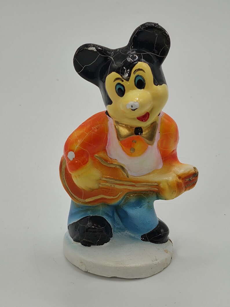 May include: A vintage figurine of a cartoon mouse playing a guitar. The mouse has a yellow face, black ears, and is wearing an orange vest, blue pants, and black shoes. The guitar is yellow and the figurine is standing on a white base.