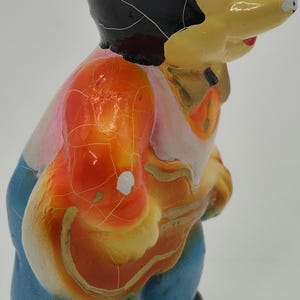May include: A vintage figurine of a cartoon character with a yellow face, black hair, and a colorful outfit. The figurine's outfit includes an orange vest, a pink shirt, and blue pants. The paint has a cracked, aged appearance.