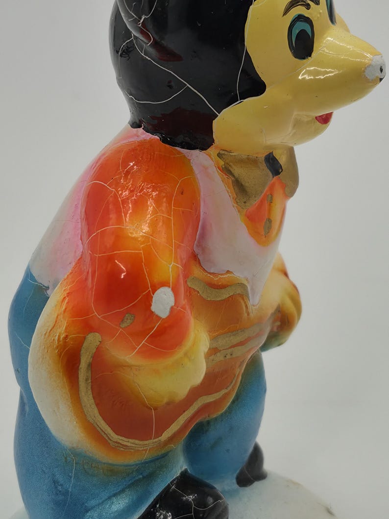 May include: A vintage figurine of a cartoon character with a yellow face, black hair, and a colorful outfit. The figurine's outfit includes an orange vest, a pink shirt, and blue pants. The paint has a cracked, aged appearance.