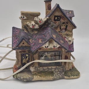 May include: A Halloween-themed ceramic house with a purple roof, adorned with glitter and small ghost figures. The house features a chimney, a window with a spiderweb design, and a stone-like base. A power cord extends from the house.