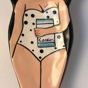 May include: A ceramic spoon rest shaped like a woman in a white swimsuit with black polka dots. She is wearing sunglasses and holding a box of cookies that says "Cookies".